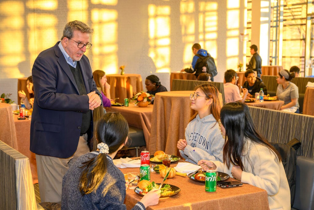 A man stands next to a Thanksgiving-themed dining table where three students are seated. The 4 people are engaged in a lively conversation. 