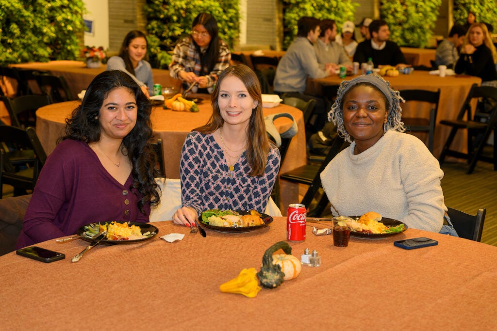 Three students sit on one side of a round table in the Thanksgiving Dinner dining hall. The table is decorated with an orange tablecloth and fall foliage center piece. A dinner plate sits in front of each student, as they pose and smile.