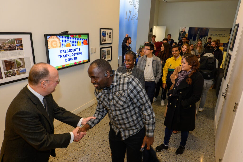 A line of students fill a hallway, waiting to be greeted by President DeGioia, who is standing at the front of the line, He smiles and shakes hands with the first student in line.