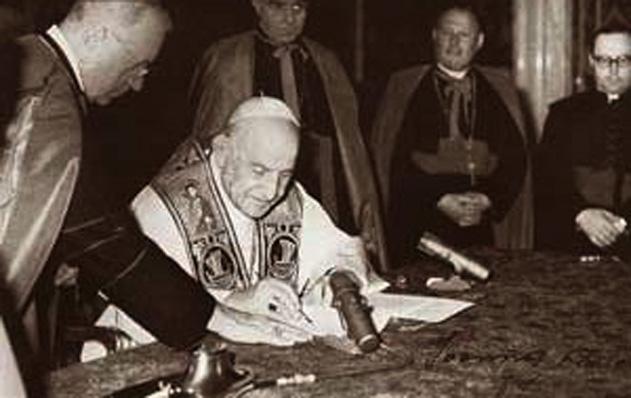 Pope John XXIII sits at a desk to sign papers.