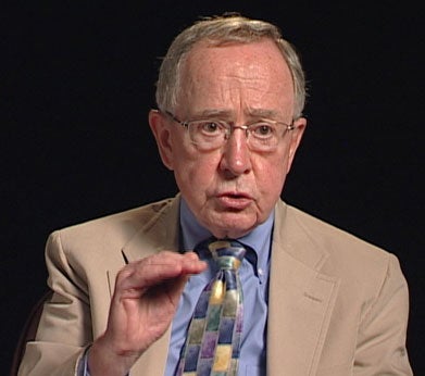 John O’Malley, S.J., a white man with greying hair and glasses, wearing a blue collared shirt and multi-colored tie, with a beige suit jacket.