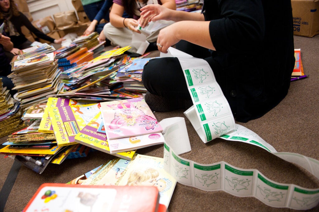 A volunteer sits on the floor among stacks of books. In their lap is a strip of book labels.