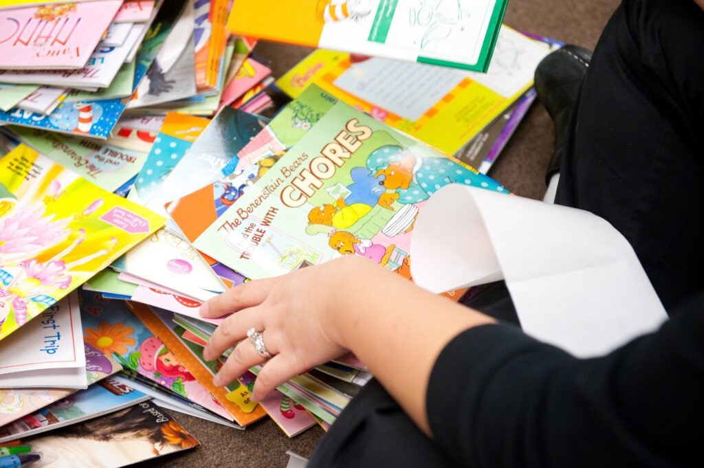 A person, whose hand is only pictured, sits in front of messy stacks of children's books.