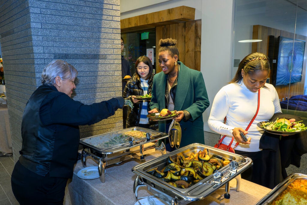 A caterer stands behind the buffet table. They serve a student food from the chafing dish that sits between them.