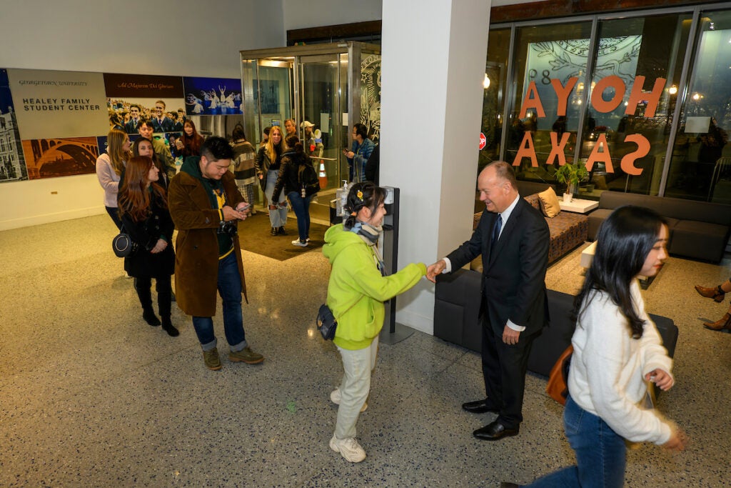 A line of students fill a hallway, waiting to be greeted by President DeGioia, who is standing at the front of the line, He smiles and shakes hands with the first student in line. The line extends around a corner, past where the camera can see.