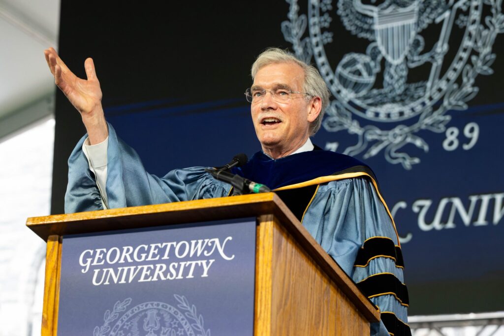 Robert Groves, a caucasian man with greying hair and a mustache, is wearing commencement robes and speaking behind a Georgetown University podium.