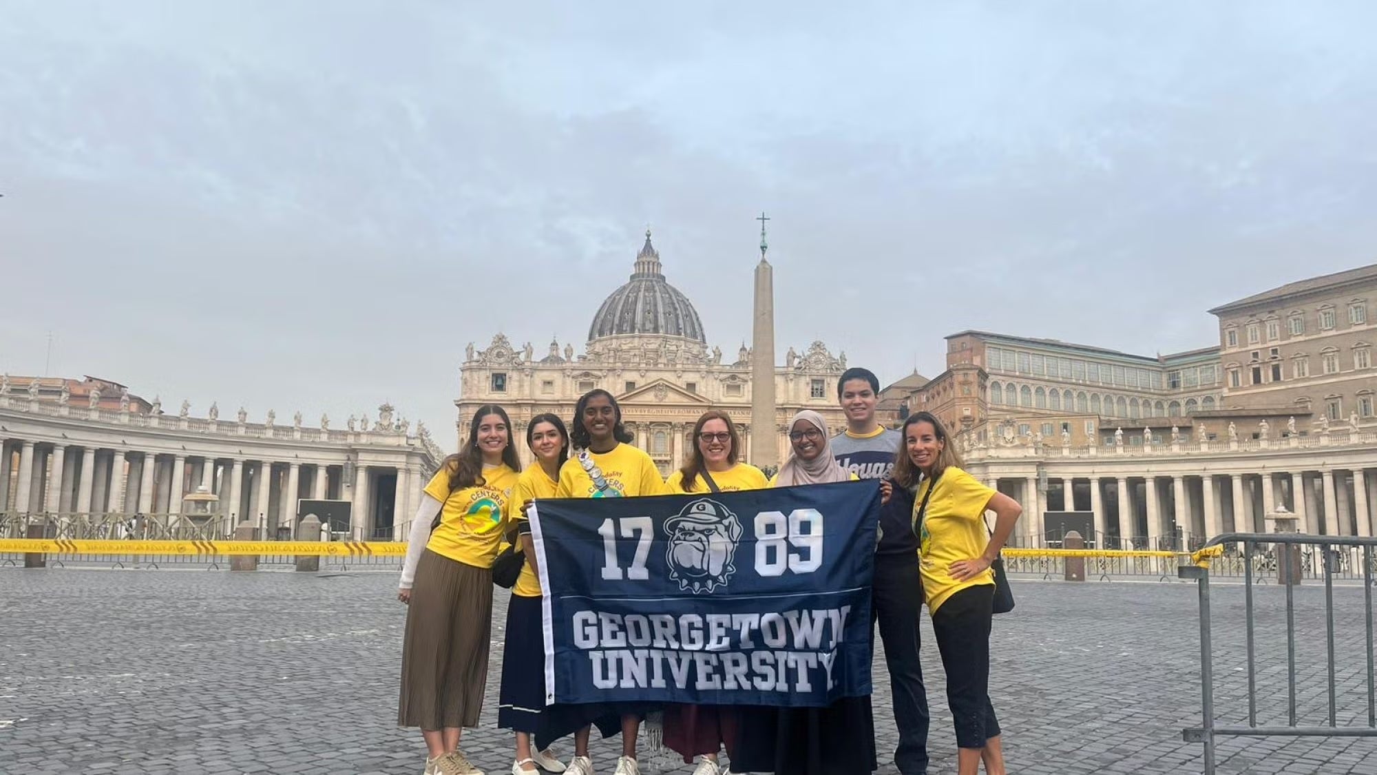 A group of seven Georgetown students hold a Georgetown University flag at the Vatican.