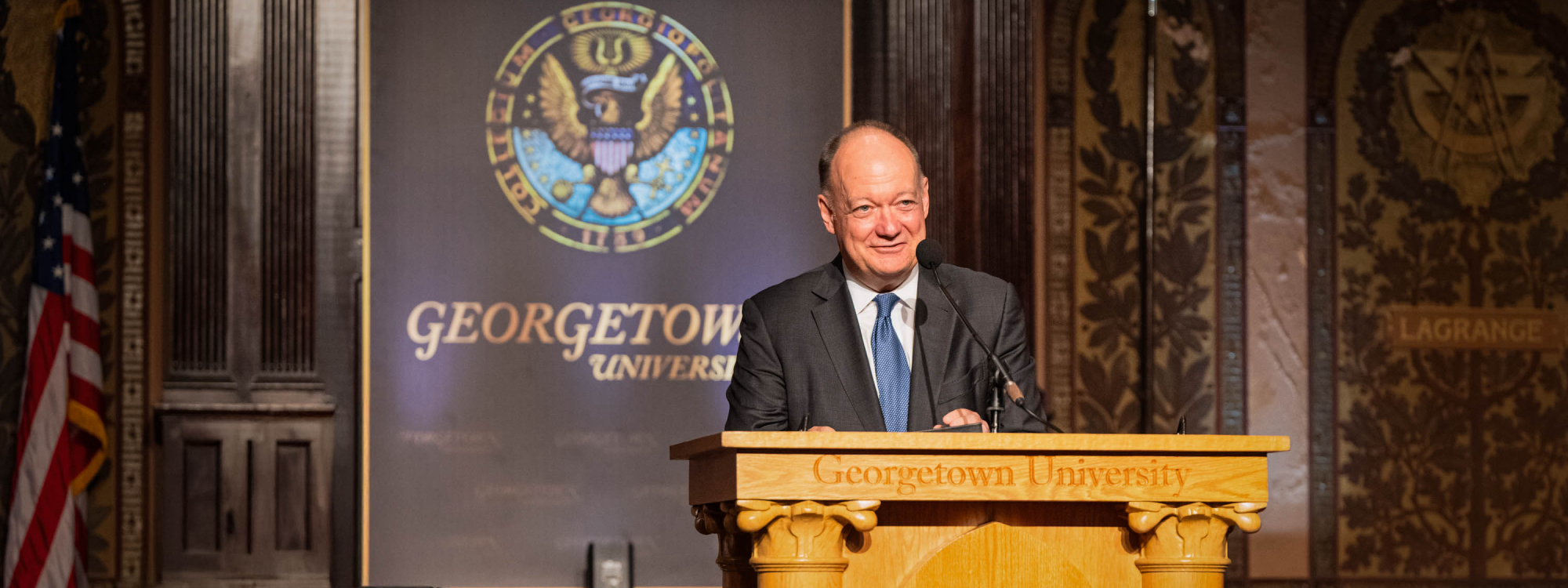 John J. DeGioia stands behind a podium, speaking to an audience. Behind him is banner with the Georgetown University name and seal.