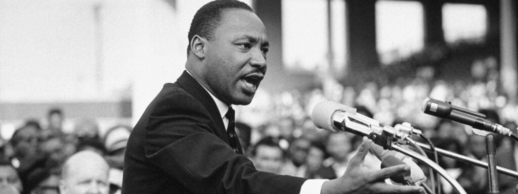 Martin Luther King stands in front of a microphone, speaking to a large crowd