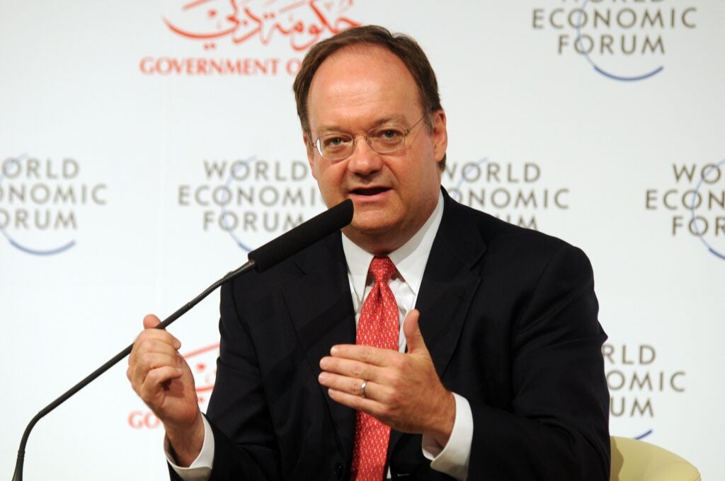 John J. DeGioia, a white man with dark hair and glasses, wearing a dark suit and red tie, speaks at a podium.