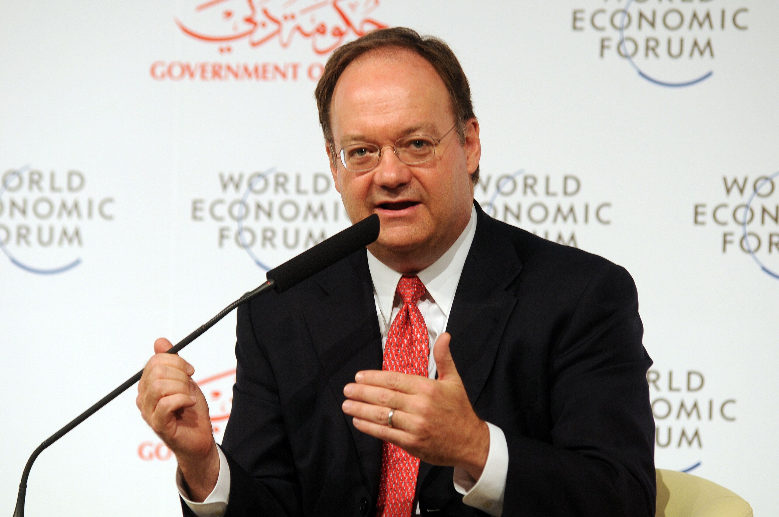 John J. DeGioia, a white man with dark hair and glasses, wearing a dark suit and red tie, speaks at a podium.