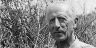 Black and white photo of Father Pierre Teilhard de Chardin, wearing a button down shirt, is standing in a field of brush.