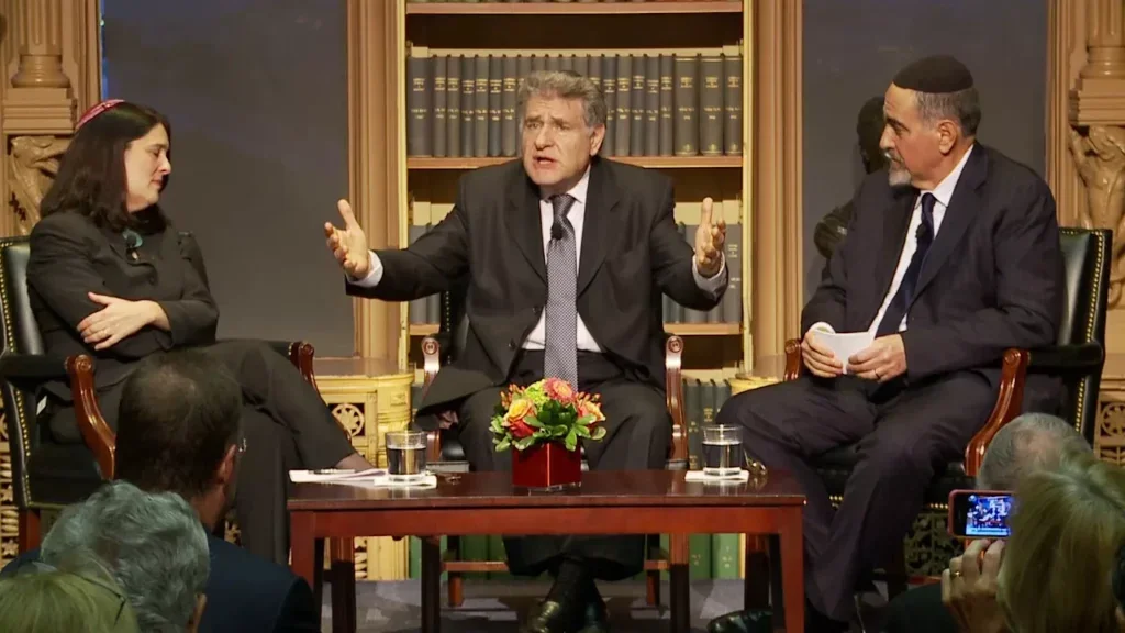 Rabbi Abraham Skorka sitting in front of a wooden bookcase, speaks to an audience. Two other panelists, a man and a woman, sit on either side of him