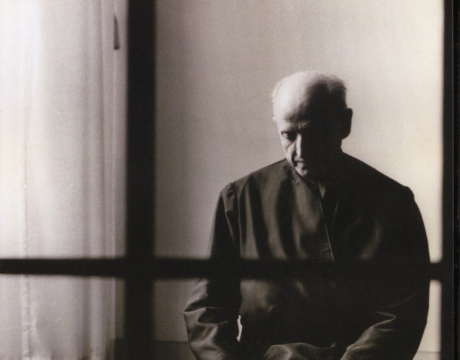 Black and white photo of Pedro Arrupe, a white balding man wearing dark robes, kneeling on the floor next to a large window.