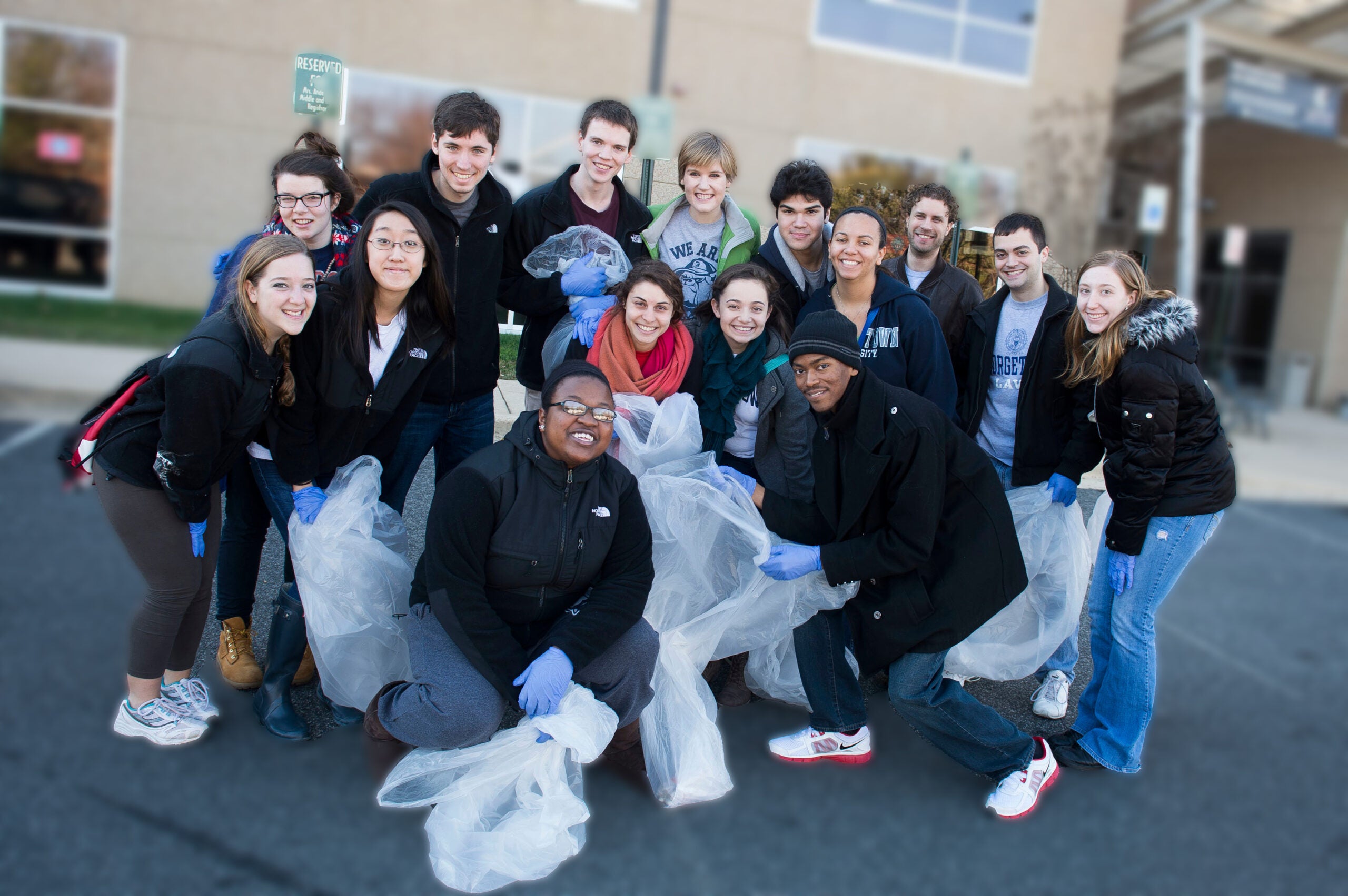 A group of 15 students pose outside a building, wearing rubber gloves and holding garbage back, prepared for a clean up service project.