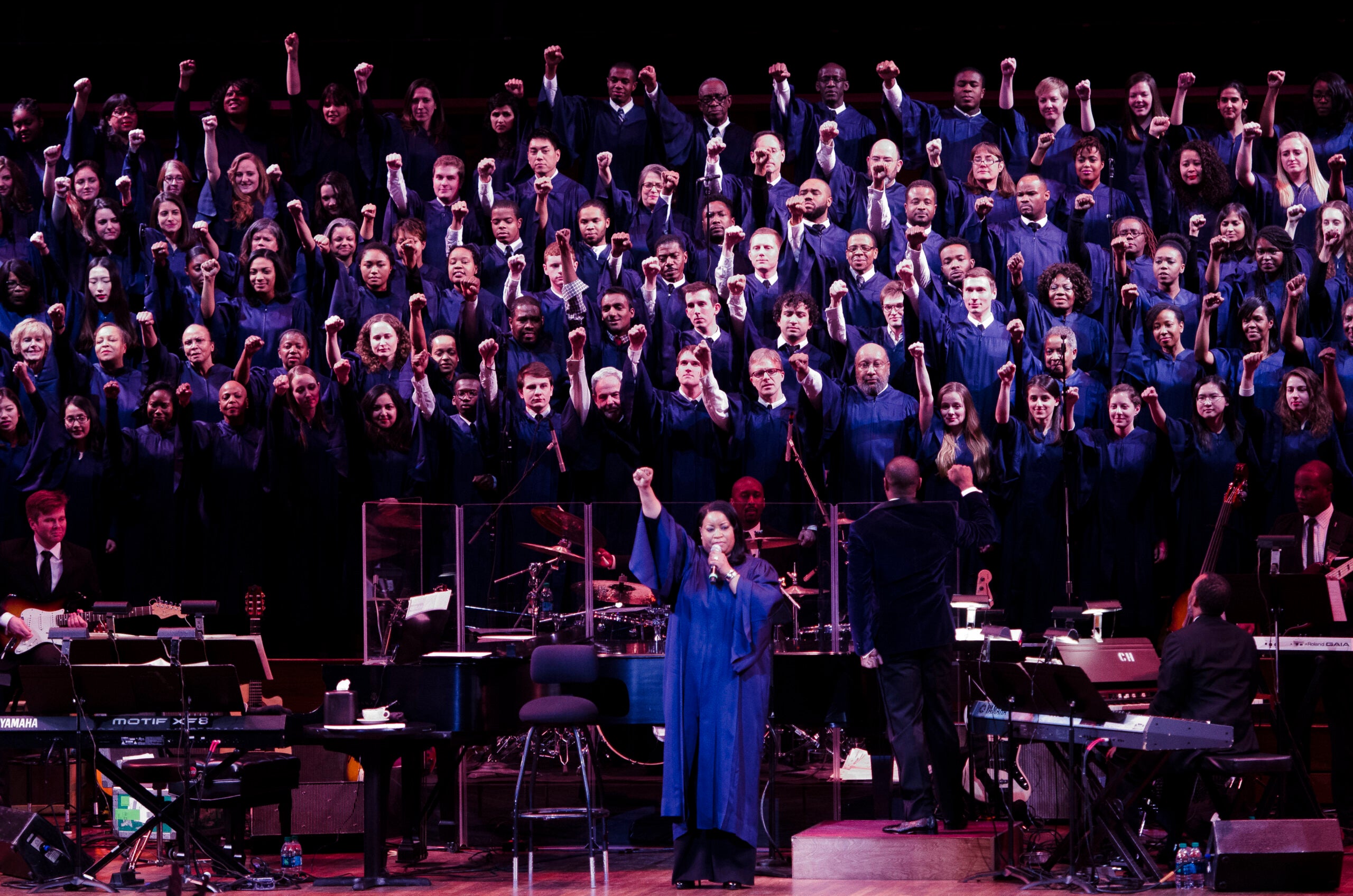 A large choir in blue robes sings, each with a fist in the air.