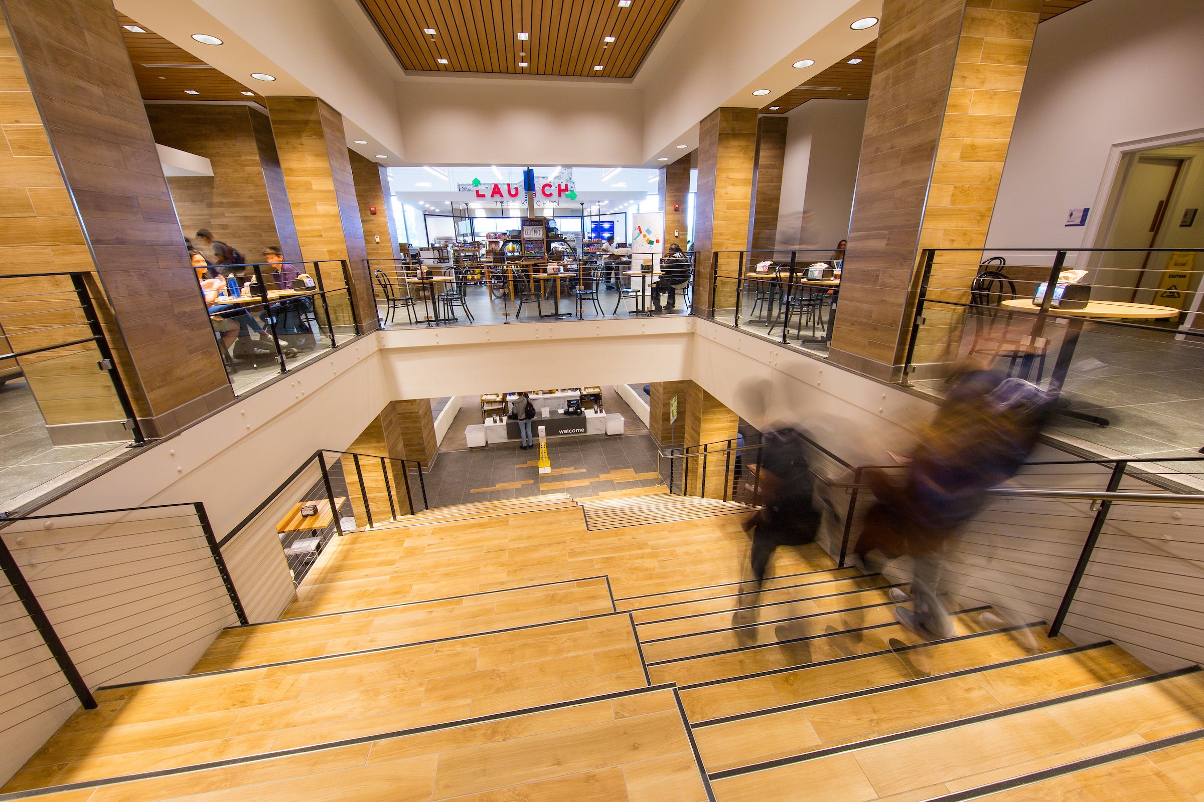 An interior open staircase in an atrium, leading down. Dining hall spaces are visible on both the top and bottom floors.