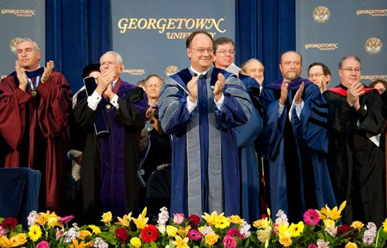 President DeGioia, wearing commencement robes, stands among a group of professors on a stage, applauding.