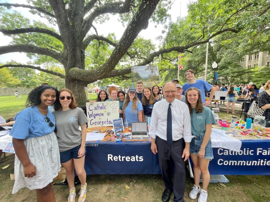 President DeGioia is outside in front of a table, posing with a large group of students.