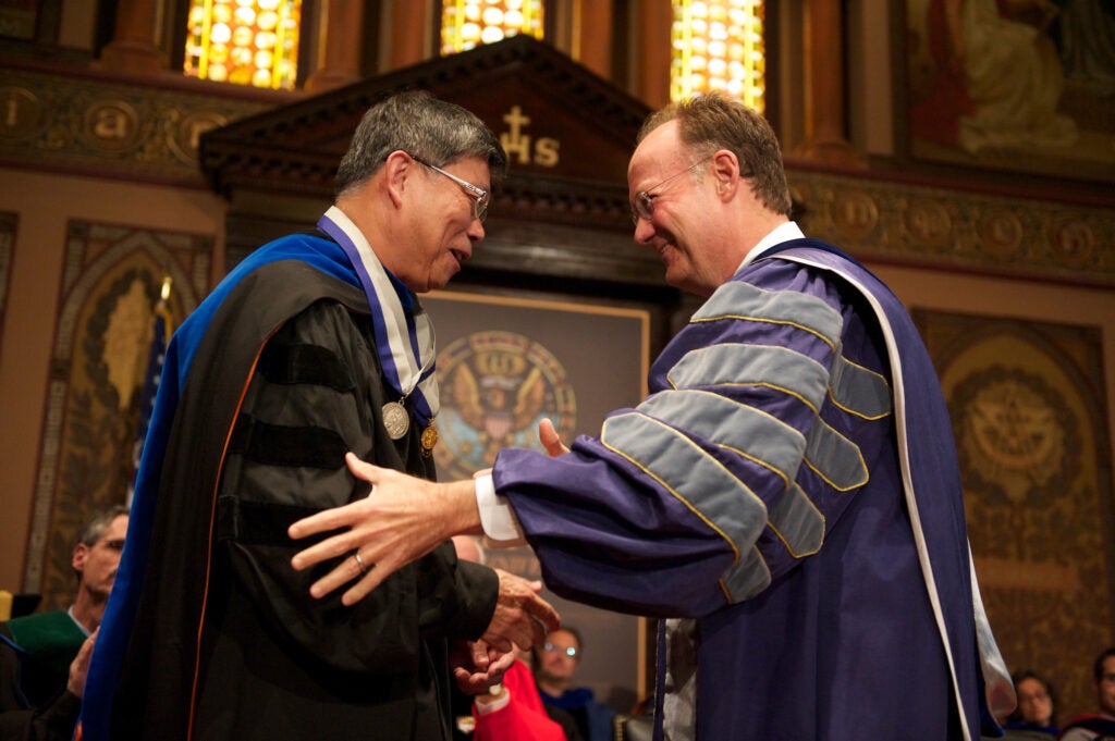Dr. Der-Chen Chang and Dr. DeGioia, dressed in academic regalia, greet each other on a stage