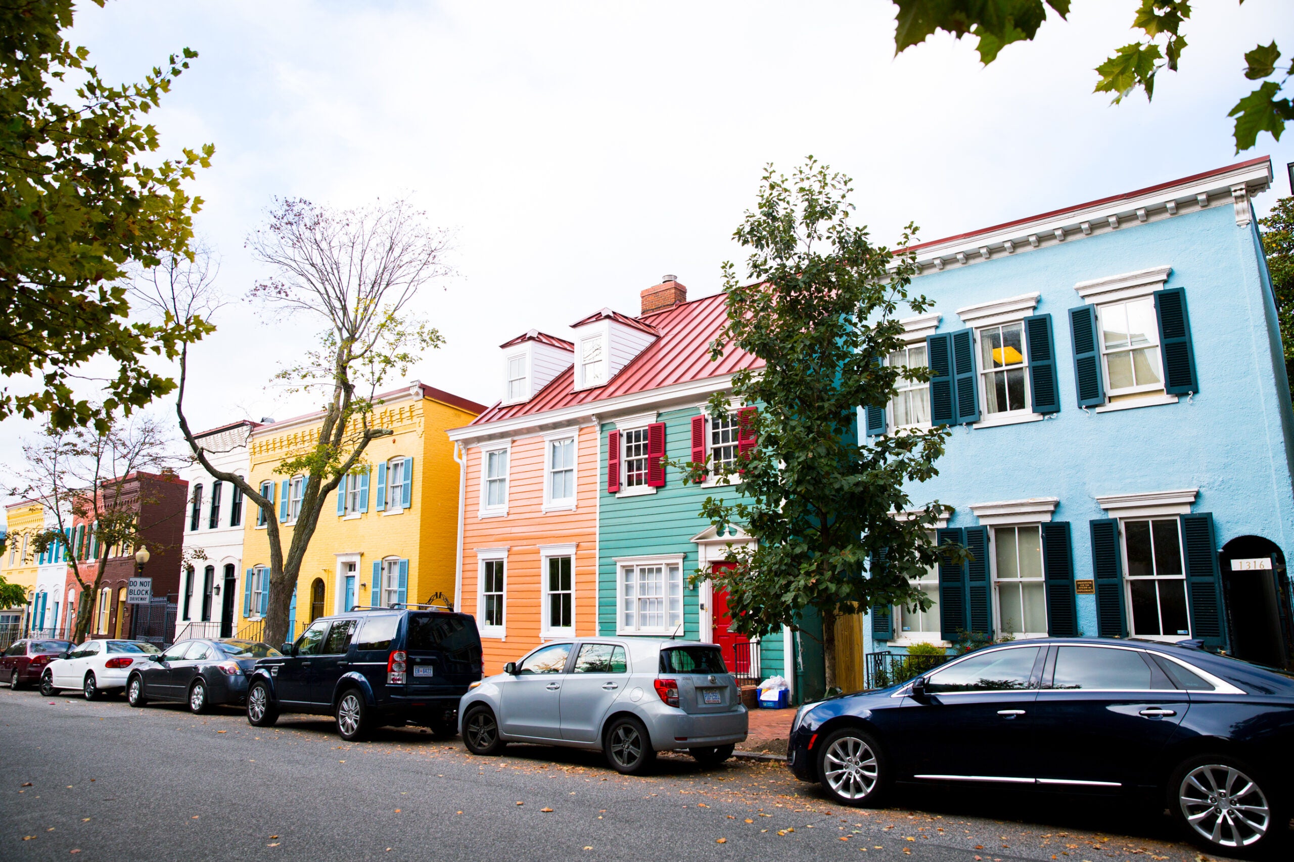 Row of colorful townhouses line a street in the Georgetown neighborhood. Cars are parallel parked along the street.