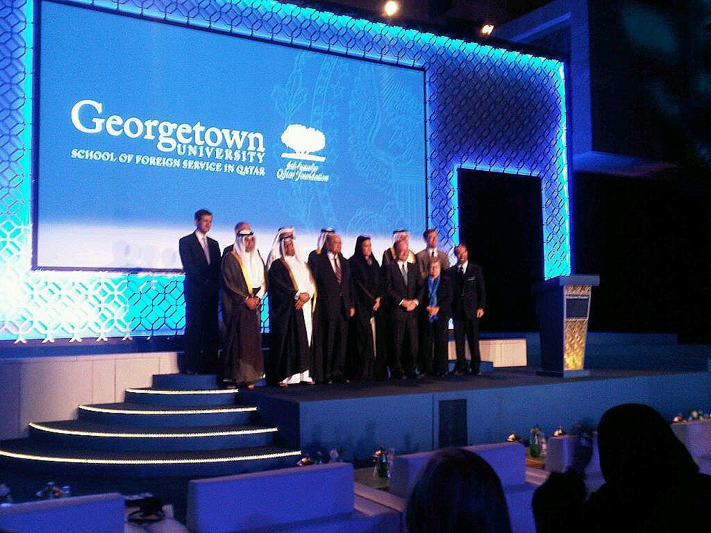 A group poses formally on a stage in front of a backdrop that reads Georgetown University School of Foreign Service in Qatar.