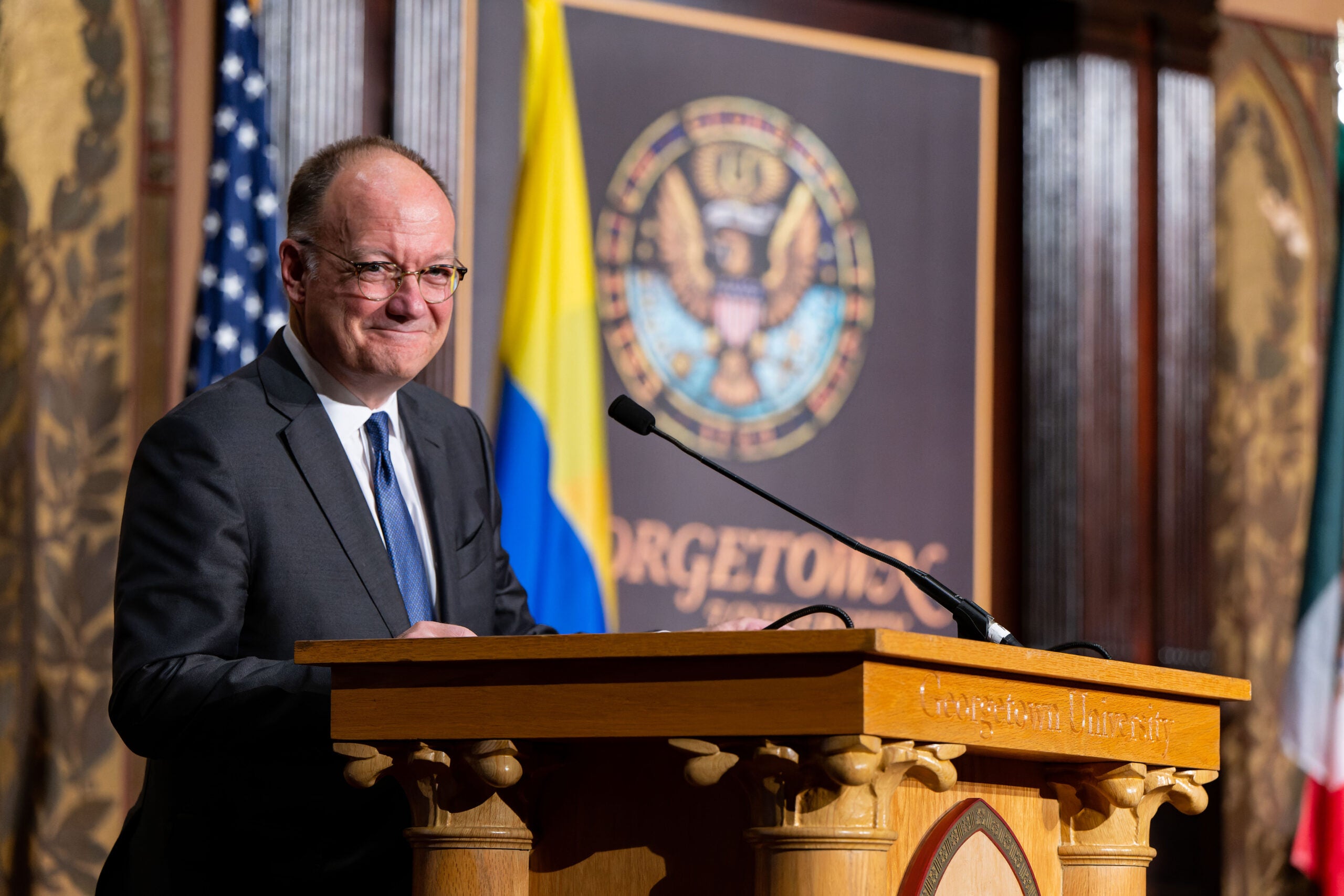 John J. DeGioia speaks at a wooden podium. Behind him is an ornate wall with the Georgetown seal.
