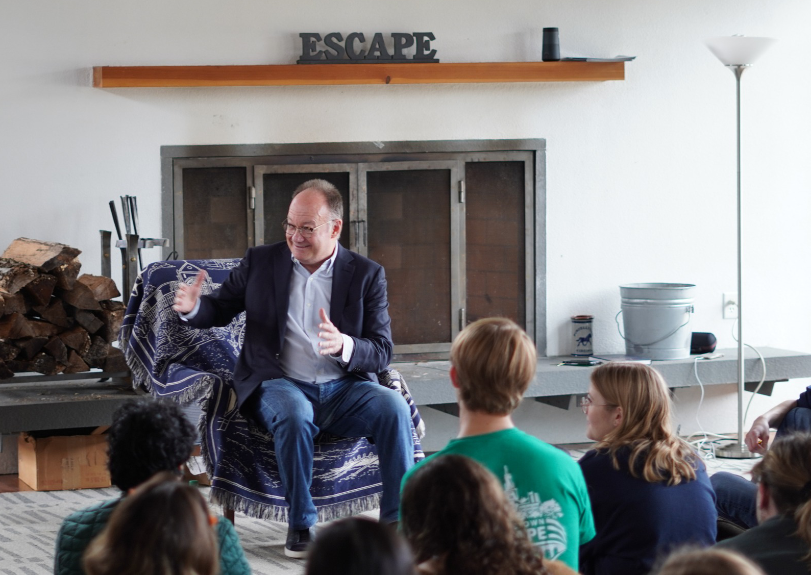 President DeGioia is seated as he speaks to a group. Behind him is a closed fireplace. The shelf above it holds a block with the word Escape on it.