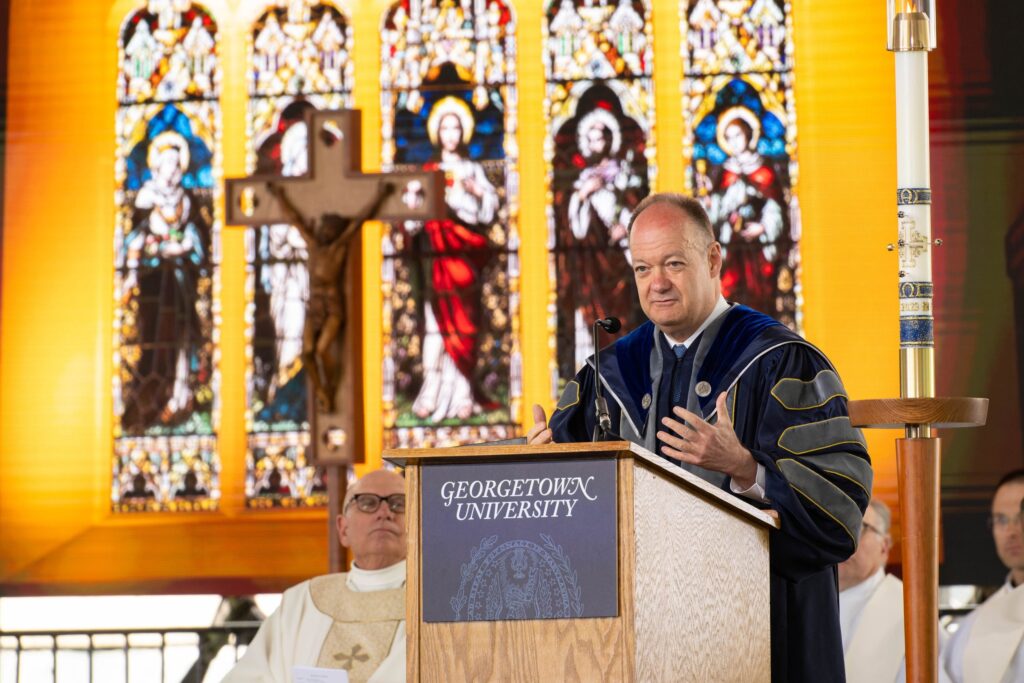 President DeGioia, wearing academic regalia, speaks from behind a podium. He is in a chapel, standing in front of a large stained glass window.