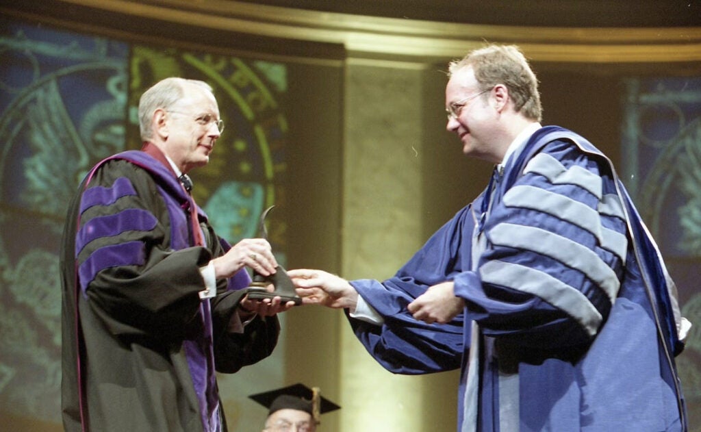 President DeGioia and Edward Quinn, both dressed in academic regalia, greet each other on a stage.