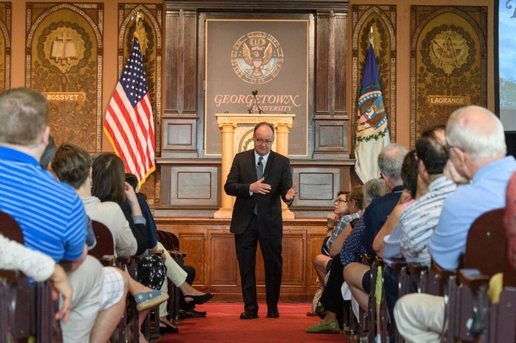 President DeGioia addresses a seated crowd in a ornately designed wood-paneled room.