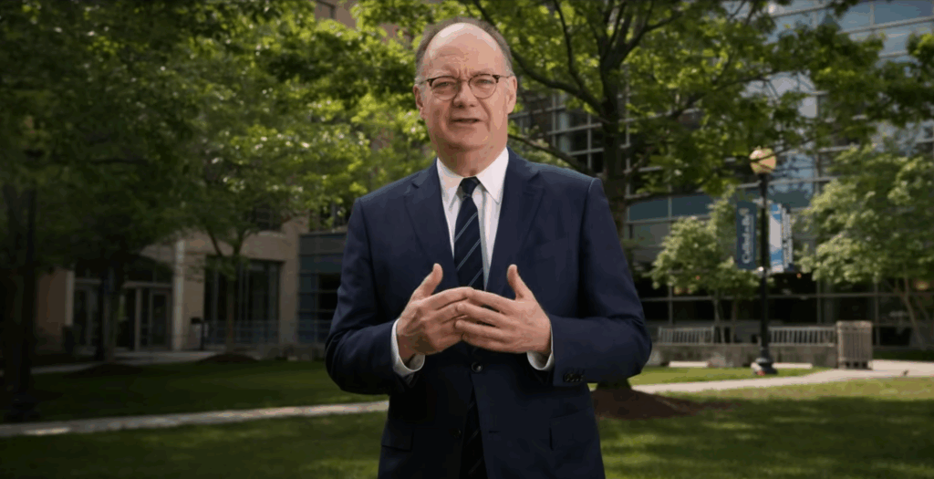 President John J. DeGioia stands on a grassy lawn outside the new construction buildings that are part of the Capitol Campus.
