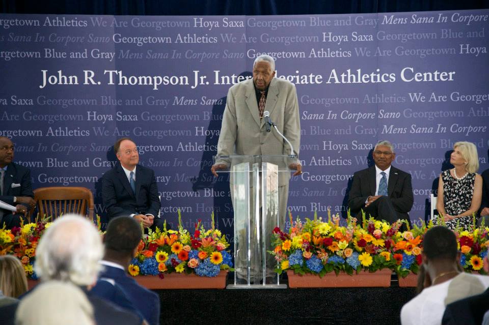 John R. Thompson speaks from behind a podium. Behind him is a wall announcing the John R. Thompson, Jr. Athletic Center. President DeGioia is seated beside him.