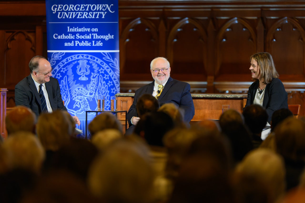 John J. DeGioia, John Carr, and Kim Daniels sit on stage in front of a Georgetown banner titled Initiative on Catholic Social Thought and Public Life.
