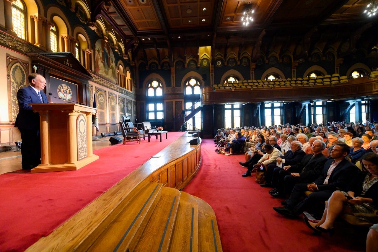 Wide shot of President DeGioia speaking on stage behind a podium to a large seated audience.