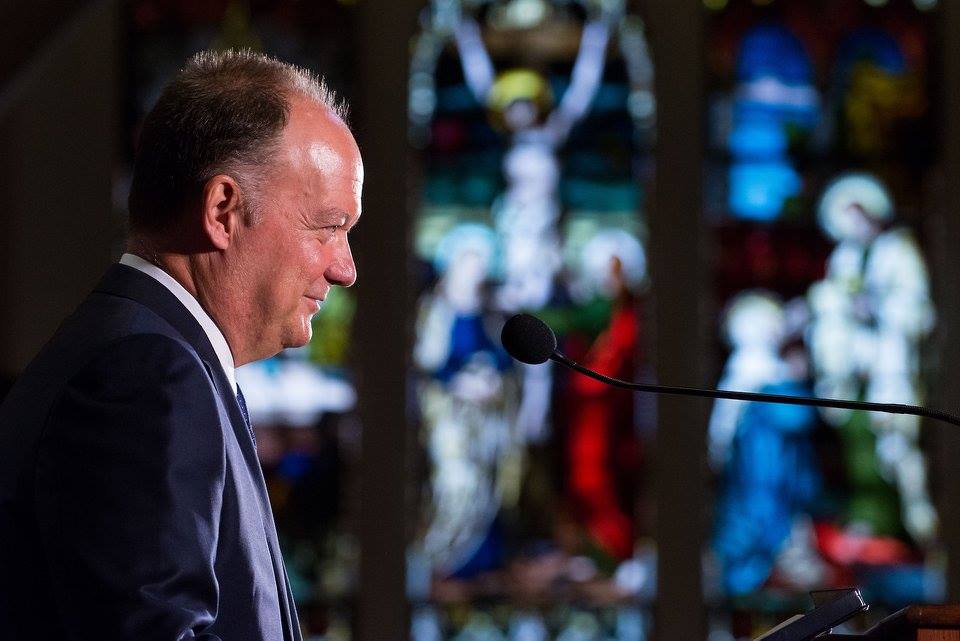 President DeGioia, standing in front of a small microphone, addresses an off-camera crowd in Dahlgren Chapel. A wall of stained glass is in the background.