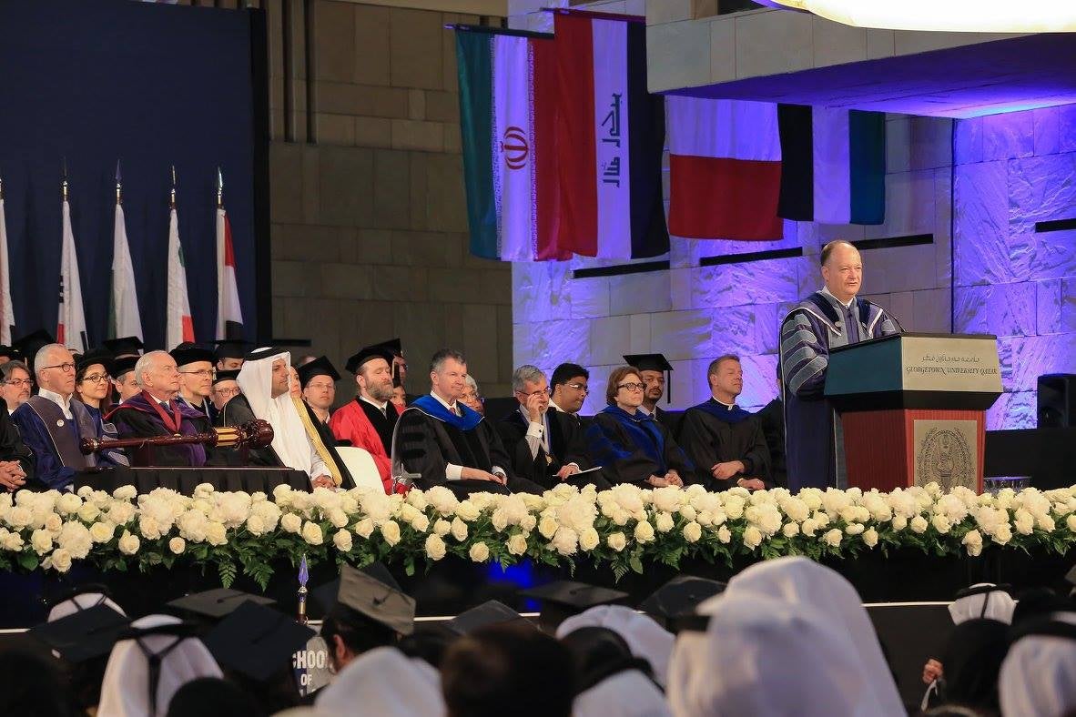 President DeGioia, wearing academic regalia speaks on stage. Behind him are seated faculty members and an array of flags from various Middle Eastern countries.