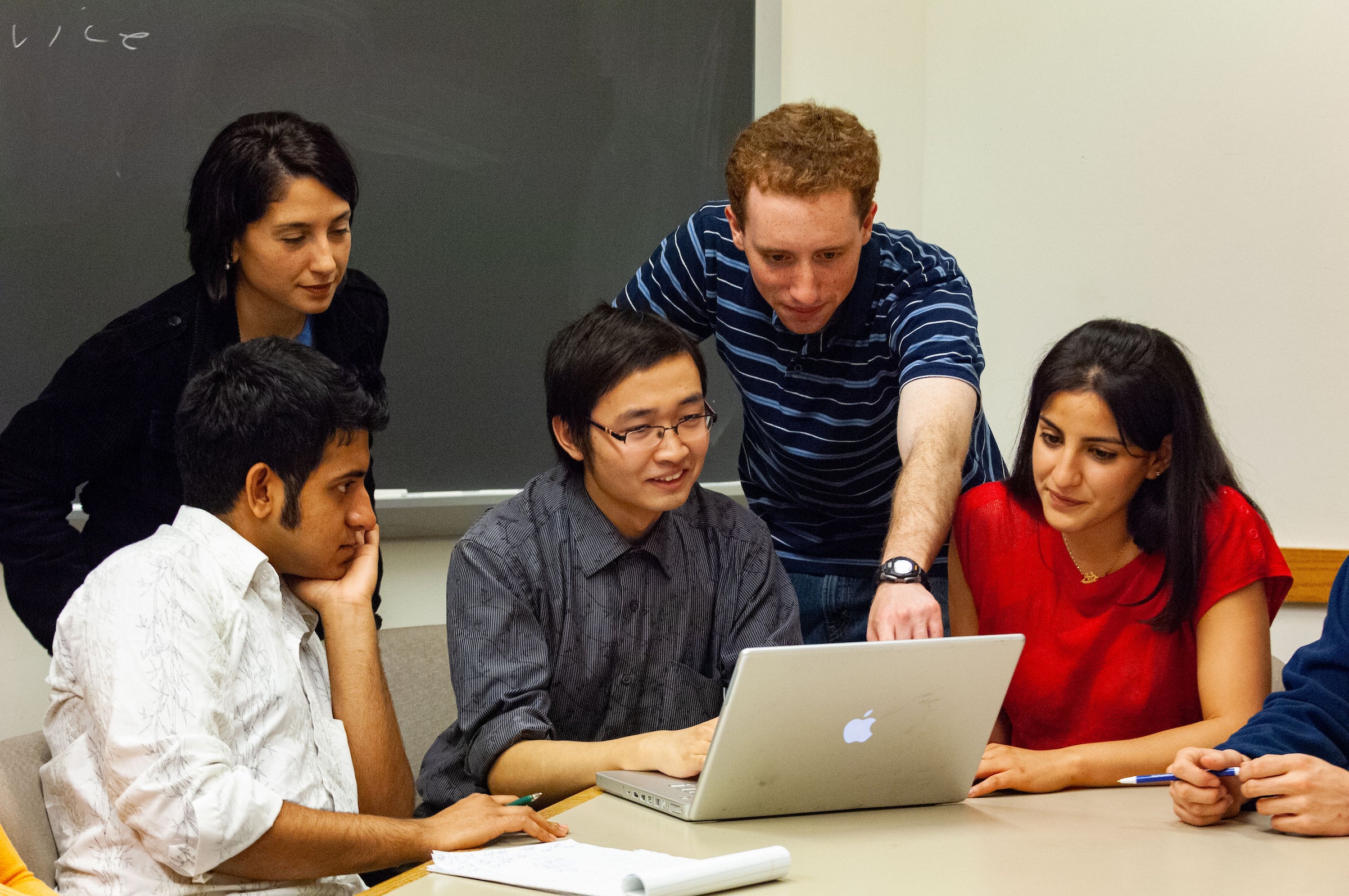 A group of 4 students gather around a 5th student and his laptop, sitting at a table in a classroom.