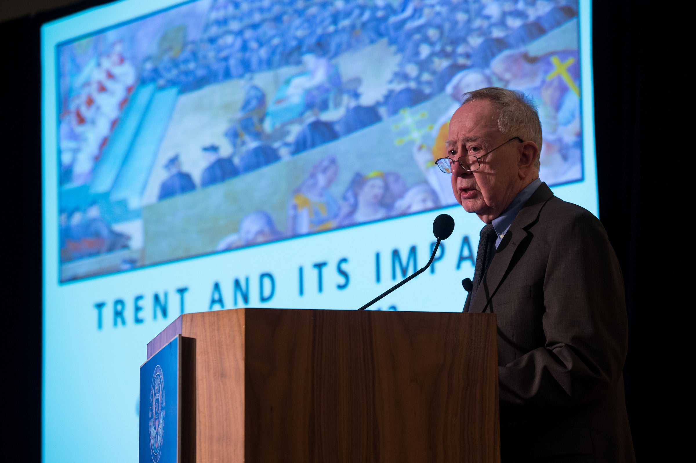 An older white man with glasses speaks at a podium. Behind him is a partially obstructed slide titled Trent and its impact.