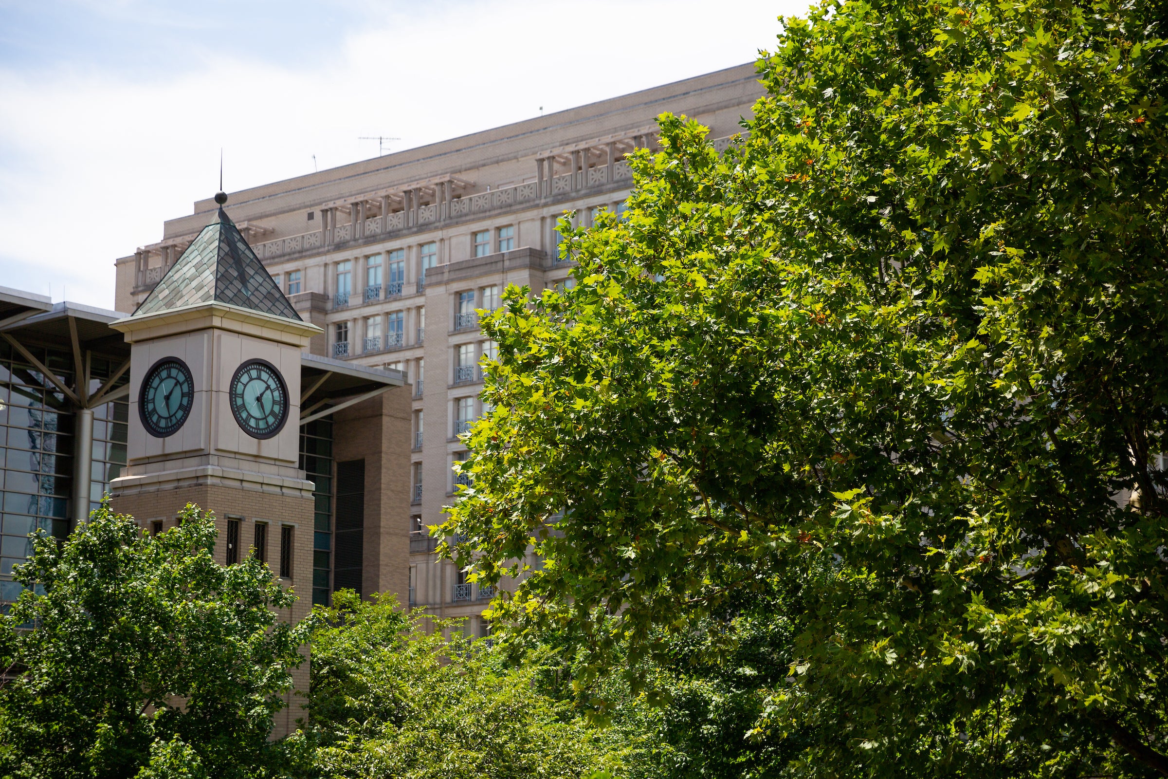 A beige clock tower framed by a large beige building in the background and trees around both.