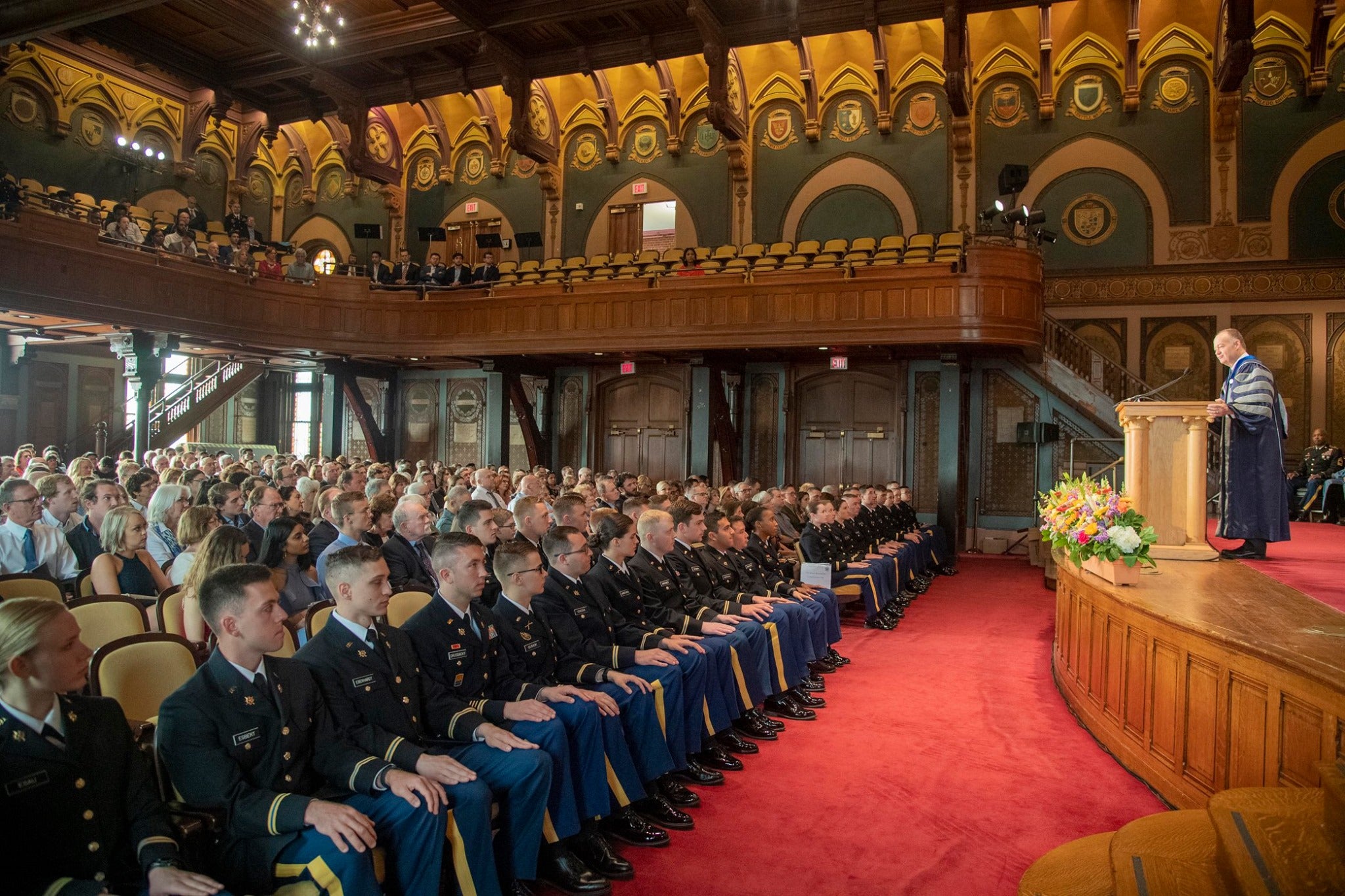 President DeGioia, wearing academic regalia, stands on a stage in front of a packed seated audience of ROTC cadets in uniform.