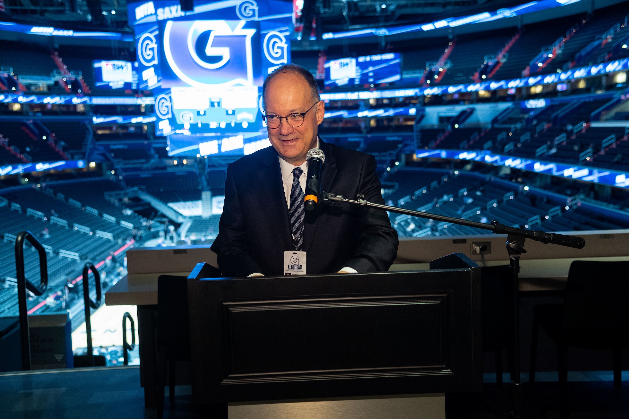 President DeGioia, wearing a suit, stands behind a podium, speaking to an audience. Behind him is an image of a Georgetown branded sports arena.