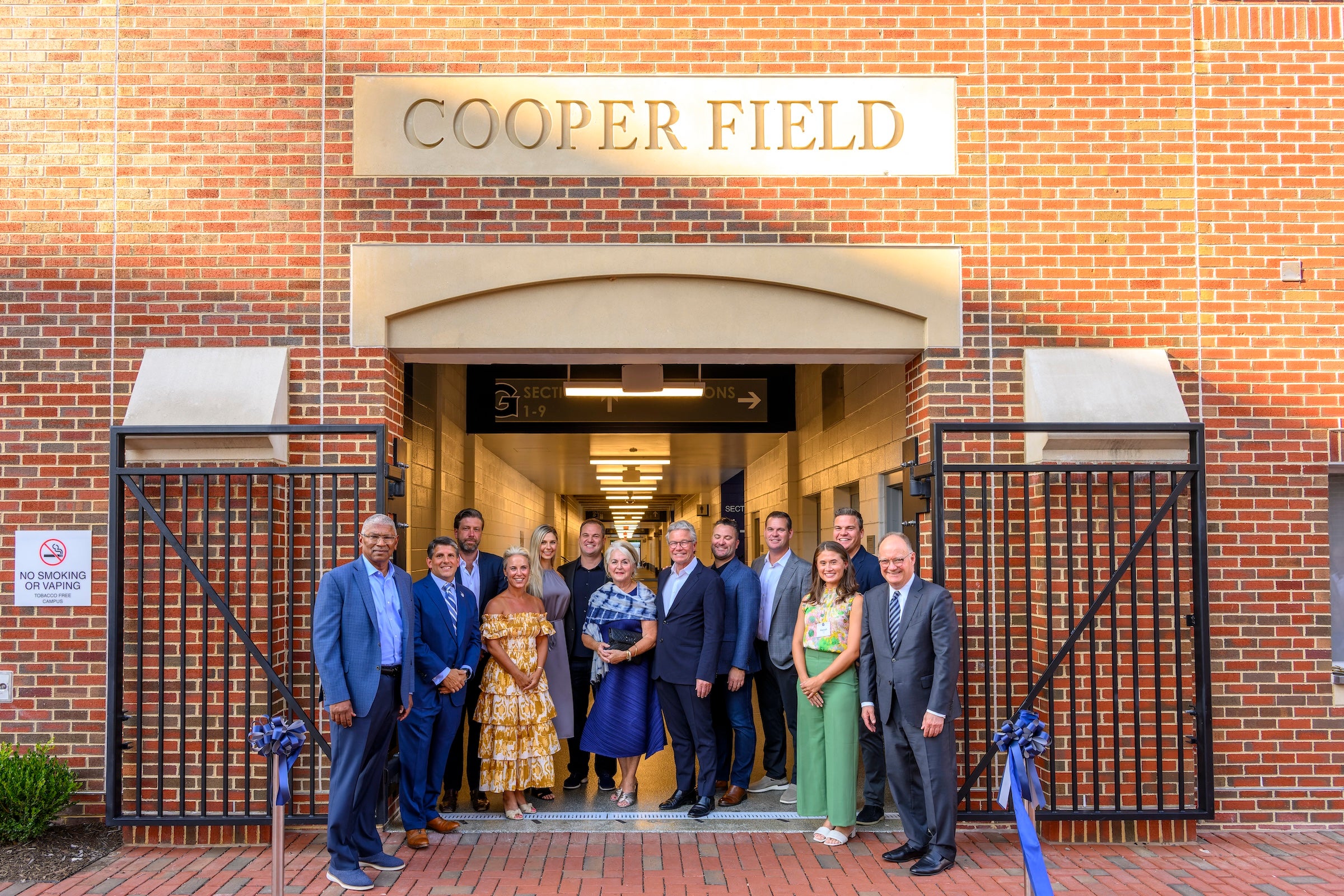 A group of 13 people dressed in formal attire stand outside the front gates of a large brick building with a stone nameplate titled 