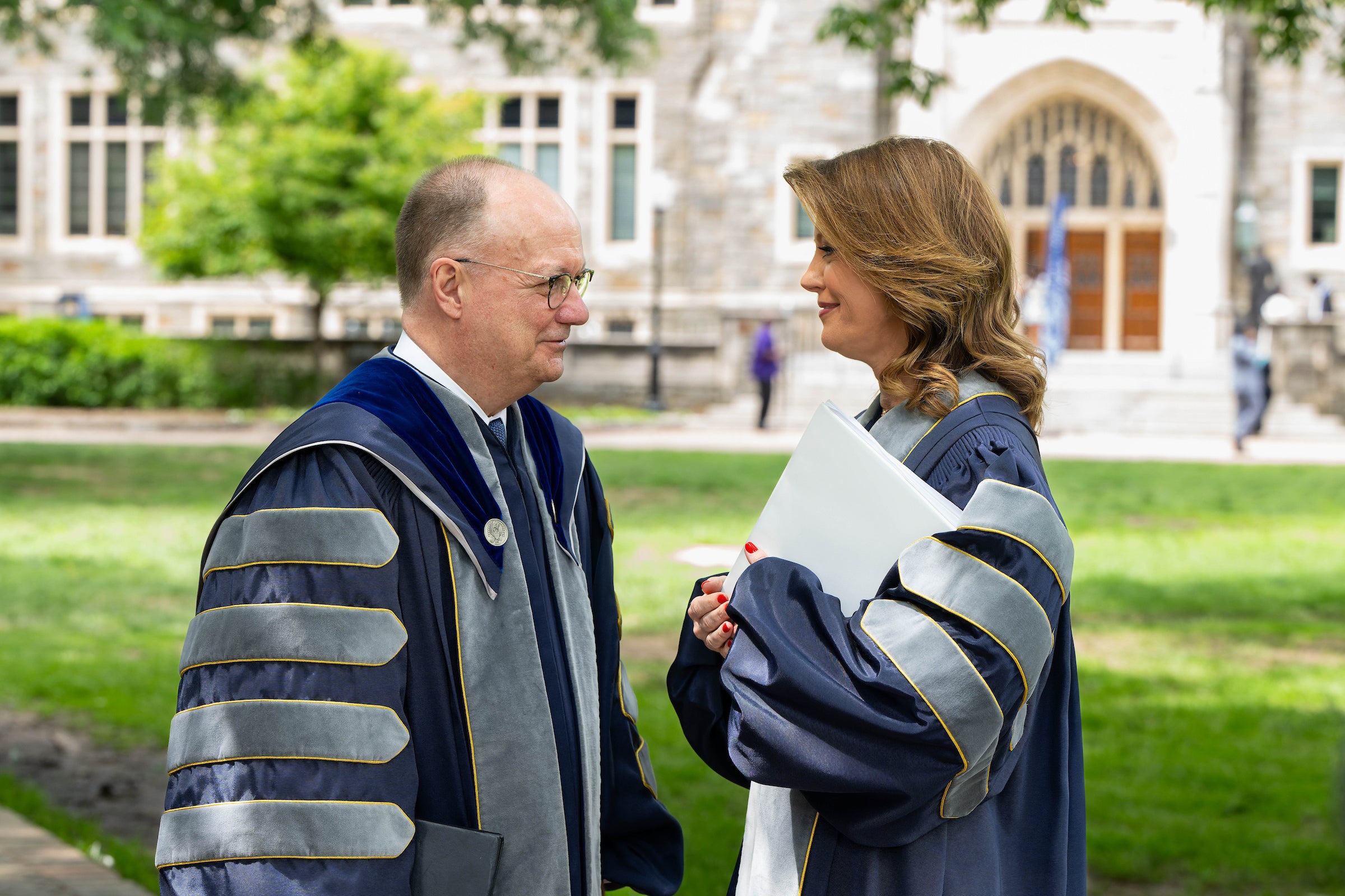 President DeGioia and Norah O’Donnell, both in academic regalia, stand facing each other in conversation on the green lawn outside Healy Hall.