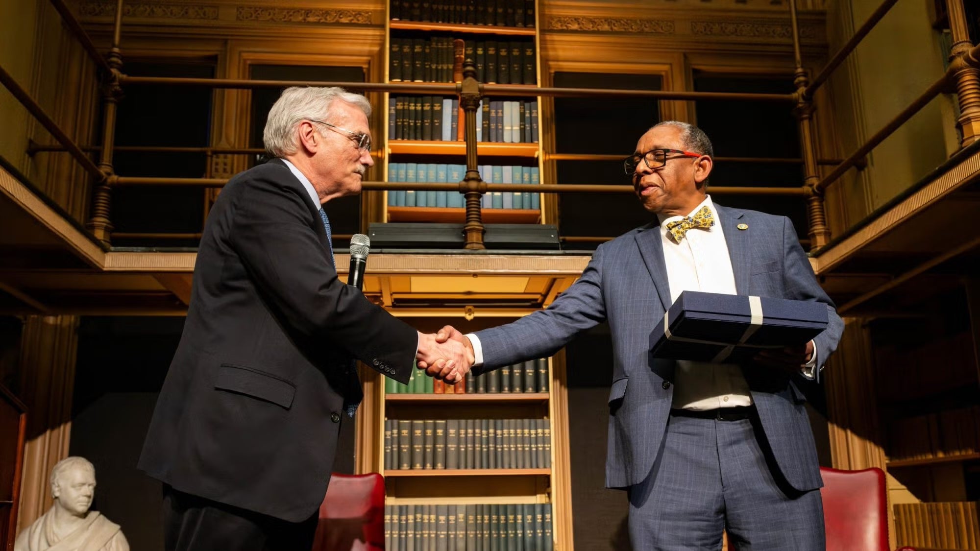 Robert Groves, a white man with grey hair, and Reynold Verret , a black man with dark hair, shake hands. Veret is holding a blue gift box. Bookshelves line the wall behind them.