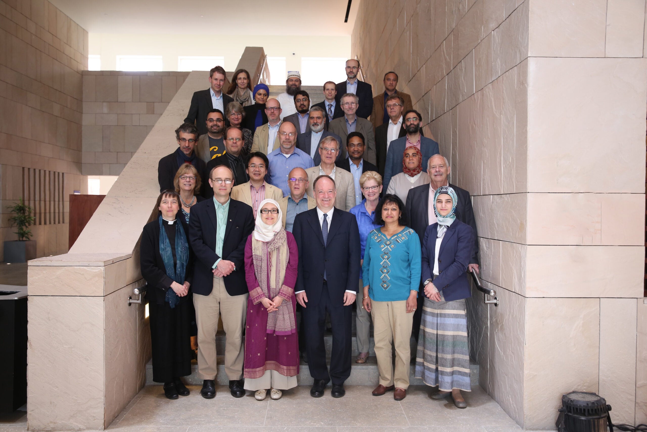 A large mixed group of people pose for a photo while standing on a half-story beige indoor staircase.