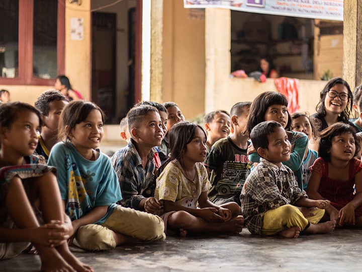 A group of seated elementary school-aged children in Indonesia look up and smile at a presentation.