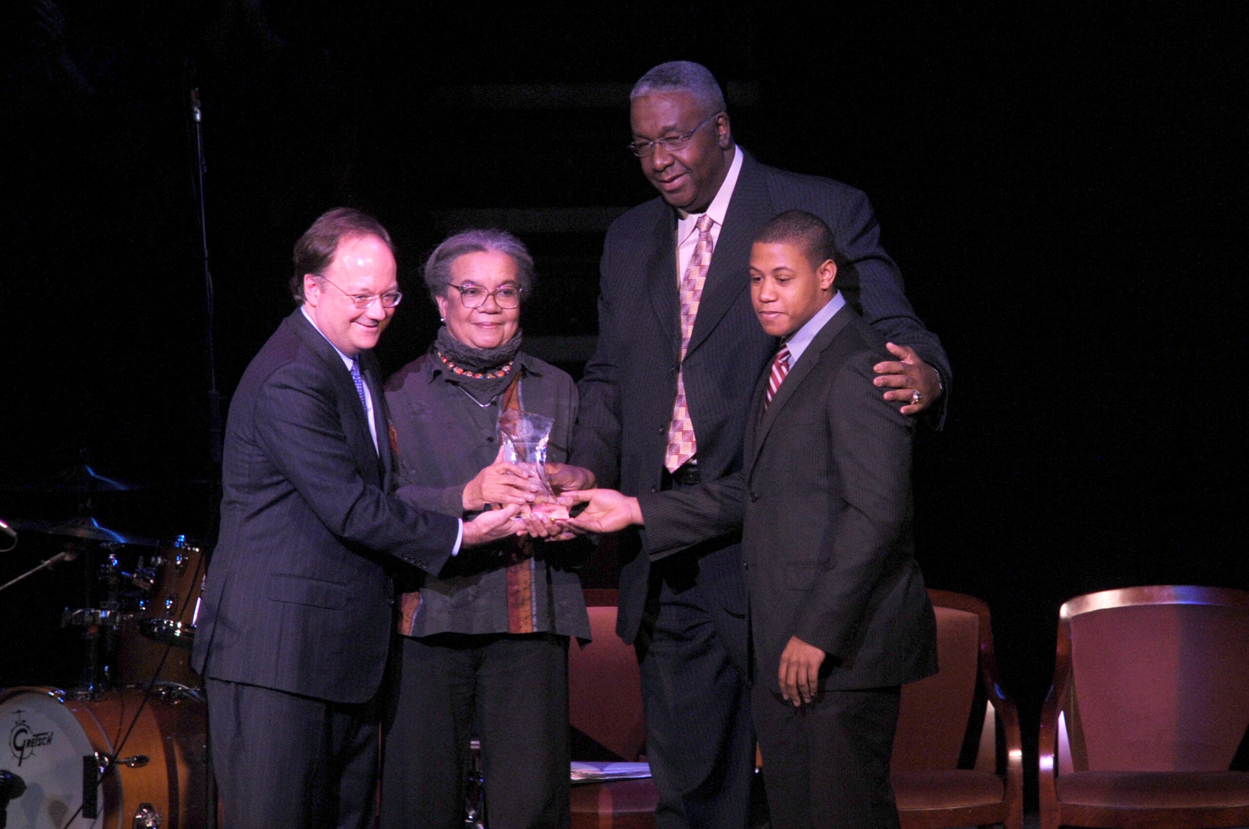 Four people stand on stage. Marian Wright Edelman stands between President DeGioia and members of the Thompson family. They all hold an award. 