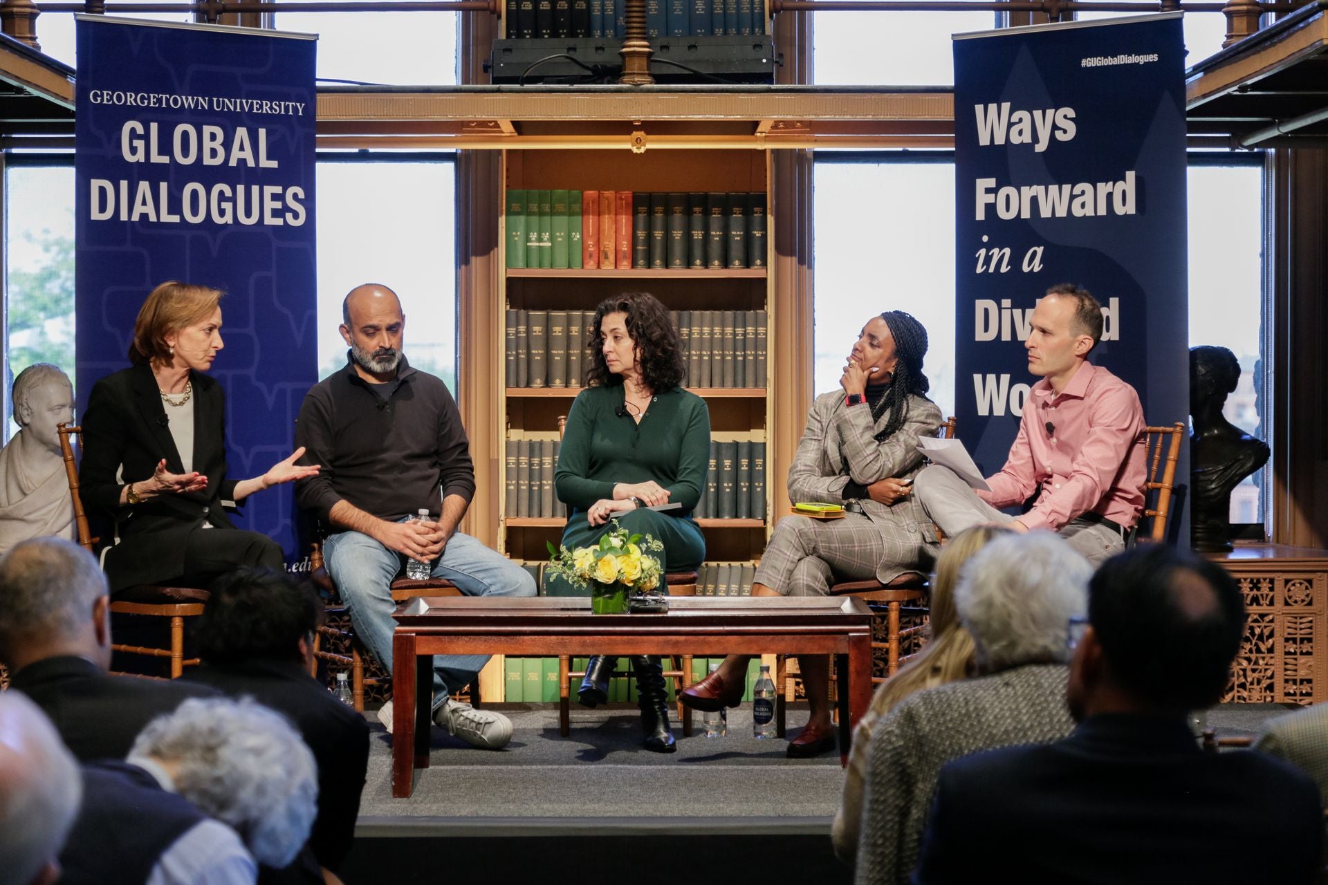 Three women and two men, all of different racial backgrounds, sit and engage in conversation, surrounded by library bookshelves.