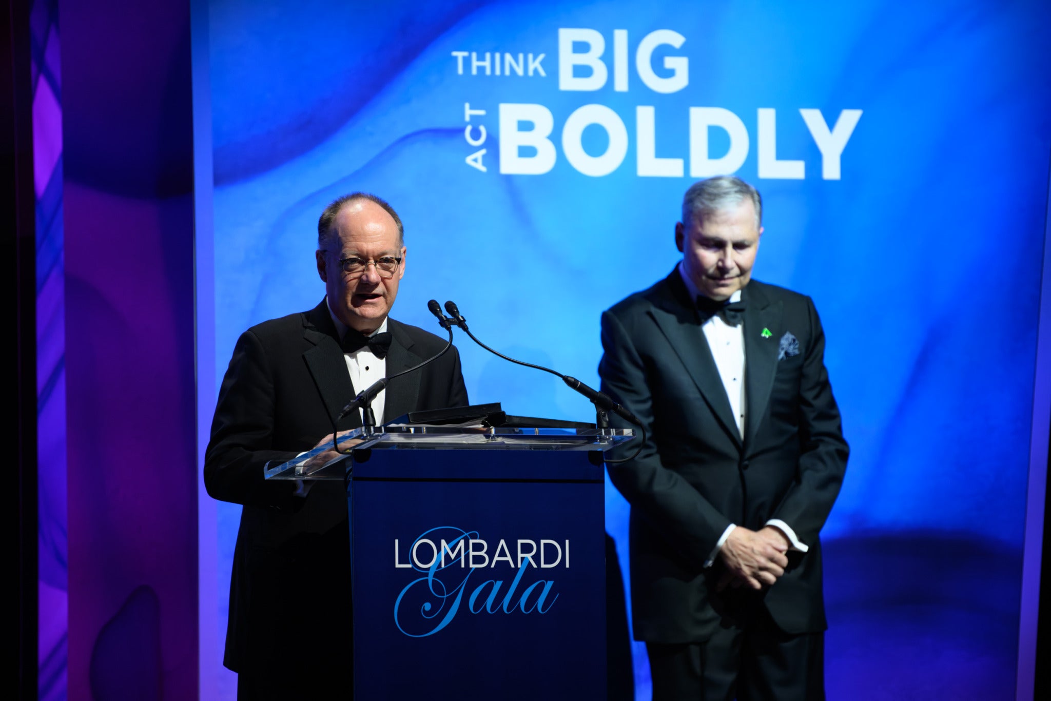 John J. DeGioia from a podium at the Lombardi Gala. Dr. Louis Weiner stands beside him. Both are wearing tuxedos. Behind them reads